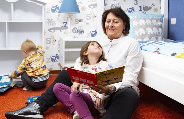  Im Borchards Rookhus liest die Mutter der Enkelin aus einem Buch vor währen die Enkelin im Schoß sitzt und hoch schaut, daneben sitz ein kleiner Junge und blättert im Buch