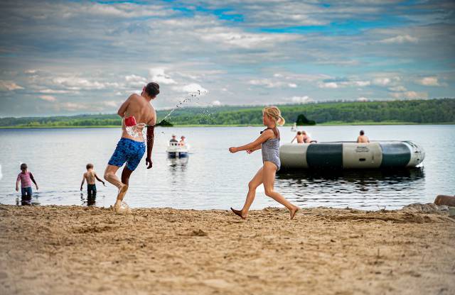 Mädchen wirft einen kleinen Eimer mit Wasser zum Papa im Sandstrand am Labussee, im Hintergrund einige Personen im Wasser