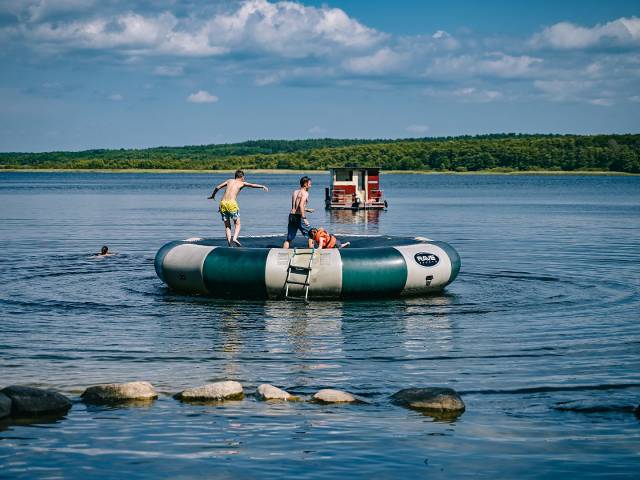 eine Familie springt auf großem Wassertrampolin im Wasser, im Hintergrund ein kleines Hausboot und eine Person schwimmt im Wasser
