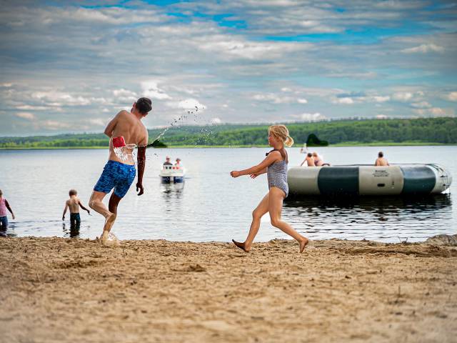 Mädchen wirft einen kleinen Eimer mit Wasser zum Papa im Sandstrand am Labussee, im Hintergrund einige Personen im Wasser