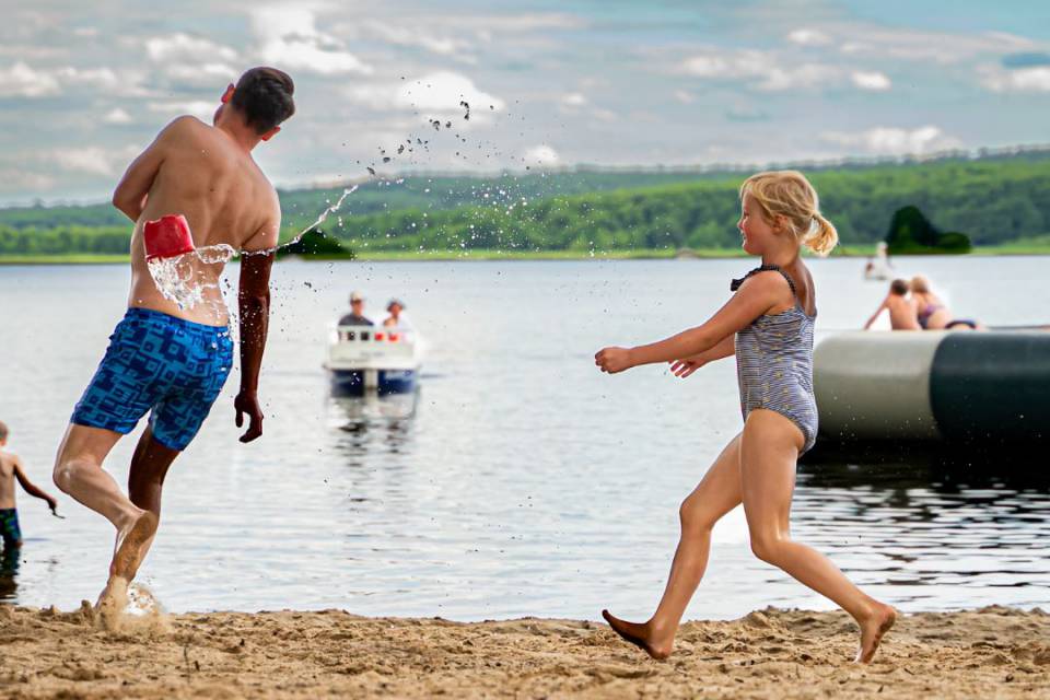 Papa und Tochter laufen über Sandstrand in Badesachen, im Hintergrund Labussee mit Personen auf dem Wasser