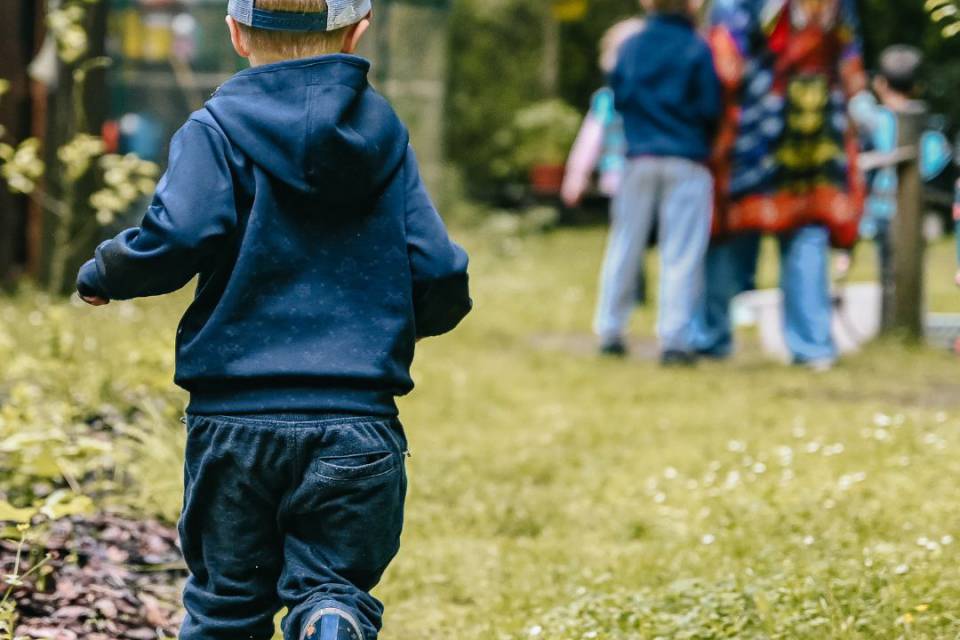 Kleiner Junge mit Kappe und Gummistiefeln läuft im Garten der Familie hinterher