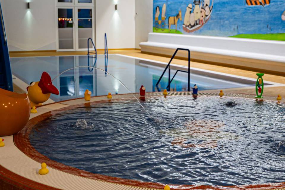 Babybecken im Schwimmbad und auf Schwimmbeckenrand stehen Quietscheenten. eine Ente spuckt einen Wasserstrahl
