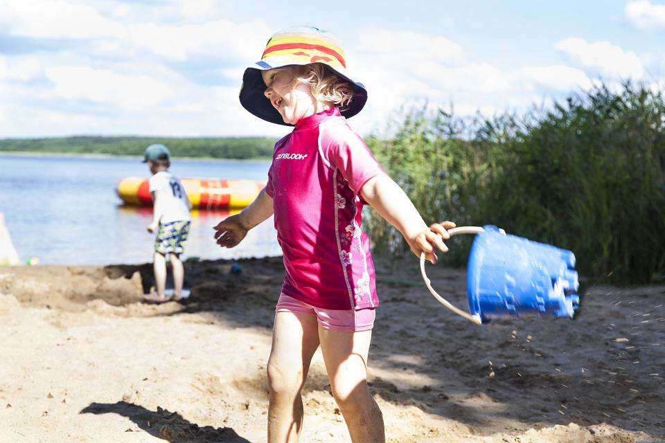 Kleines Mädchen mit Hut und Schwimmkleidung, blauem Eimer in der Hand lacht am Strand