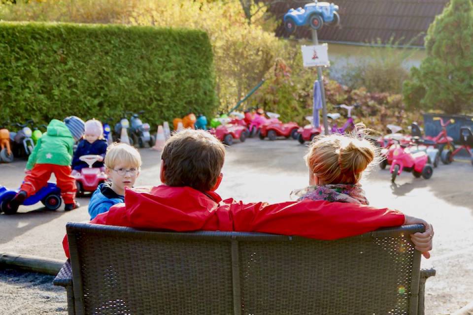 Eltern sitzen in der Sonne und schaue den Kindern beim Bobbycar fahren zu