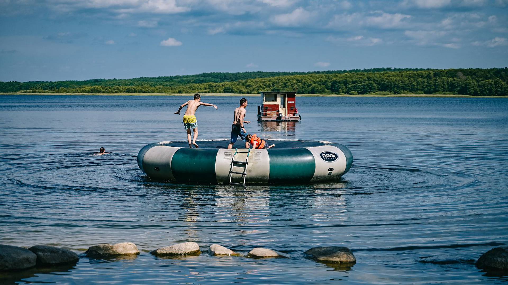 eine Familie springt auf großem Wassertrampolin im Wasser, im Hintergrund ein kleines Hausboot und eine Person schwimmt im Wasser