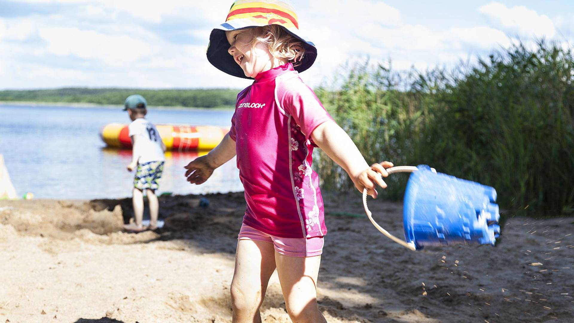 Kleines Mädchen mit Hut und Schwimmkleidung, blauem Eimer in der Hand lacht am Strand