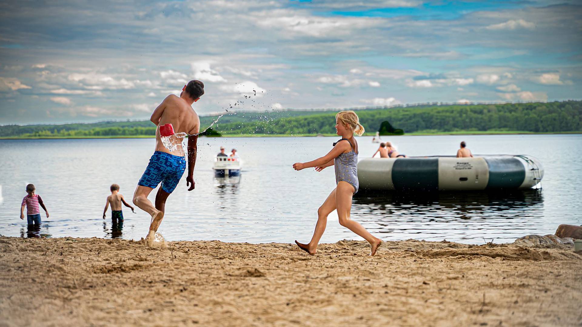 Mädchen wirft einen kleinen Eimer mit Wasser zum Papa im Sandstrand am Labussee, im Hintergrund einige Personen im Wasser